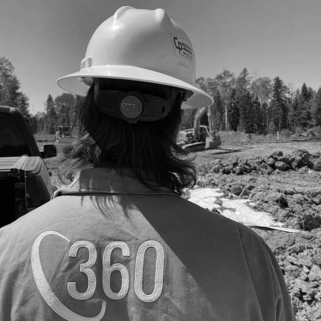The back view of a 360 employee in full protective gear and sporting a mullet. The photo was taken at a job site so there is environmental reclamation equipment in the background and the employee is observing the progress of the work.