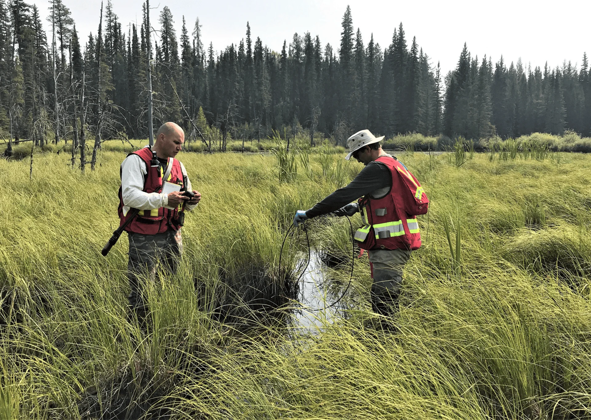 360 employees conducting water analysis during a spill response project. Photo shows 2 people standing in a flooded, grassy area holding water analysis instruments and wearing protective clothing.